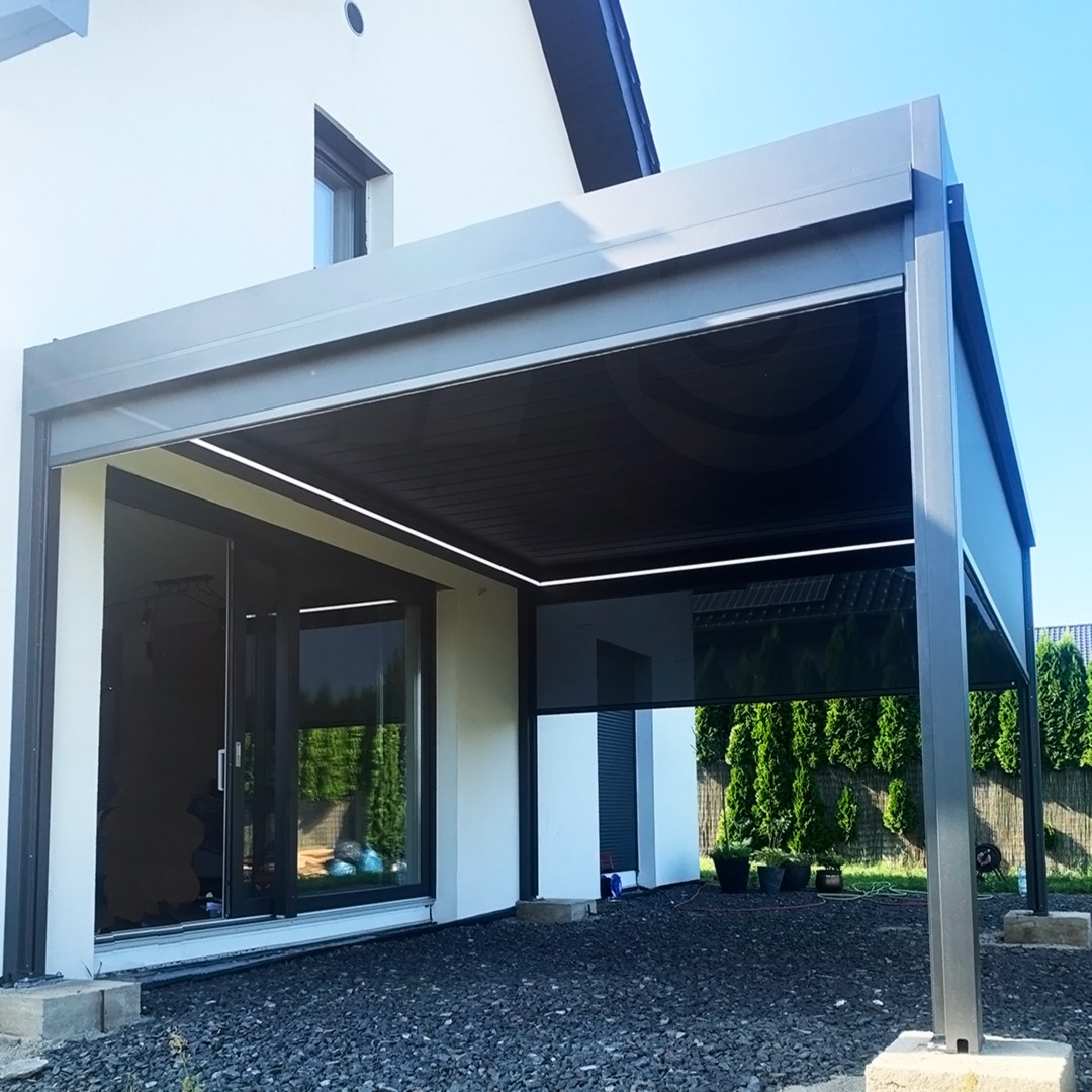 Nowoczesna, szara konstrukcja z metalu, przypominająca zadaszenie tarasu, z oświetleniem LED i częściowo zwiniętą, zaciemniającą roletą, przylegająca do białego budynku.