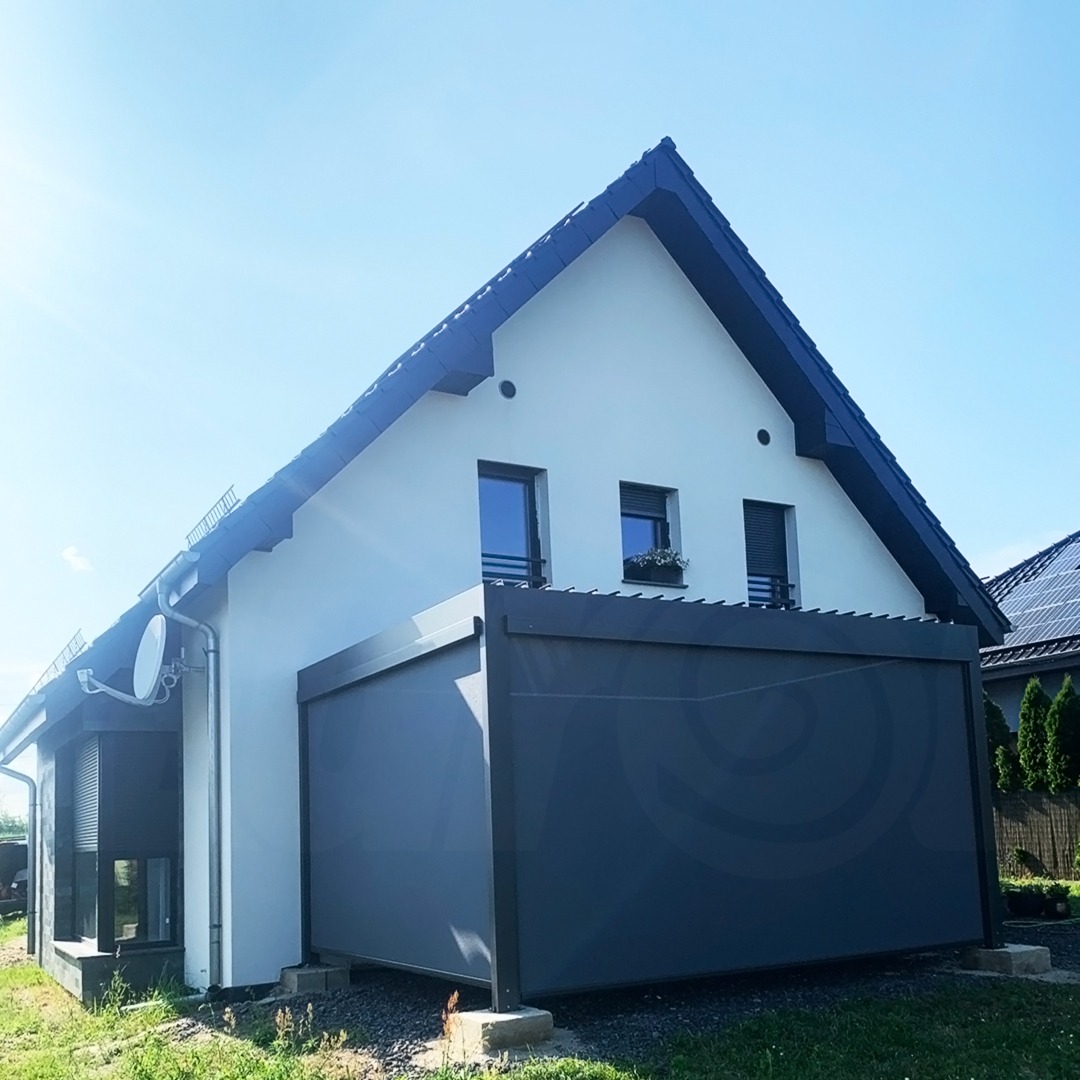 Nowoczesny dom jednorodzinny z szarą pergolą z rozwijanym bokiem, zapewniającą cień. Widoczny fragment rolety zewnętrznej na oknie. Słoneczny dzień.