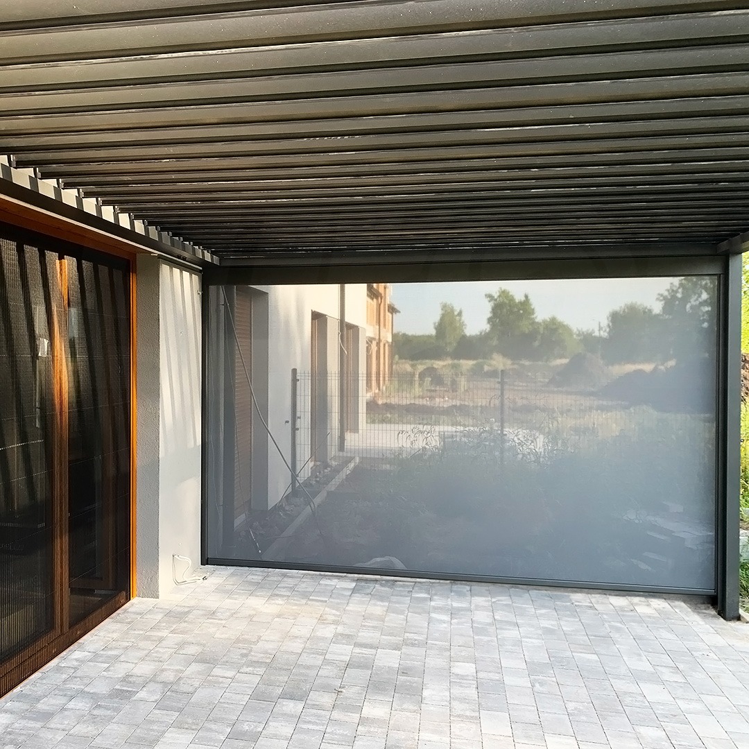 Zadaszony taras z metalową konstrukcją, osłonięty boczną, zwijaną przesłoną, widok na ogród.