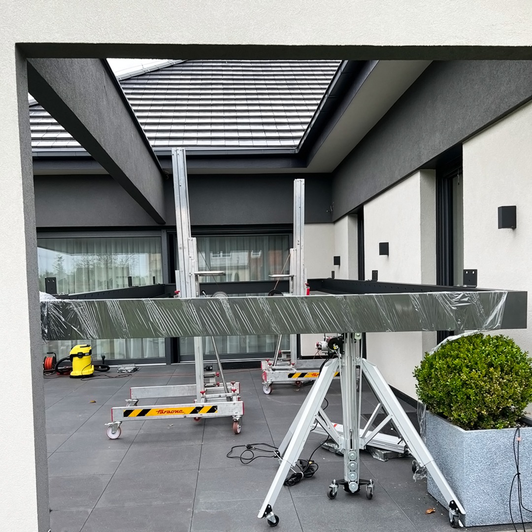 Montaż elementu konstrukcyjnego z użyciem podnośników na kółkach, widoczny fragment nowoczesnego domu z oknami i oświetleniem zewnętrznym.