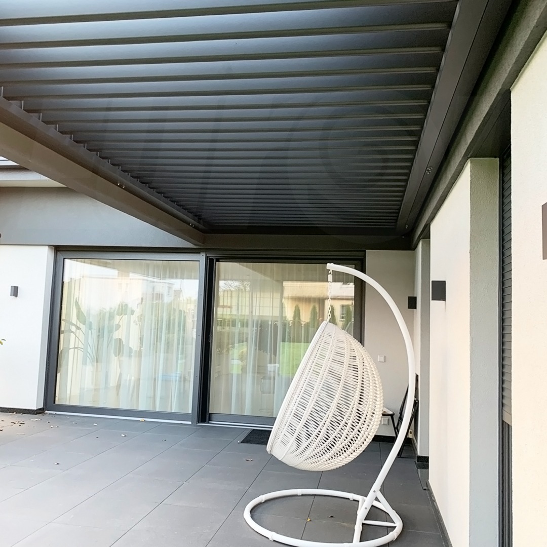 Nowoczesny taras z metalową pergolą w kolorze antracytowym i wiszącym fotelem kokon z białego rattanu.
