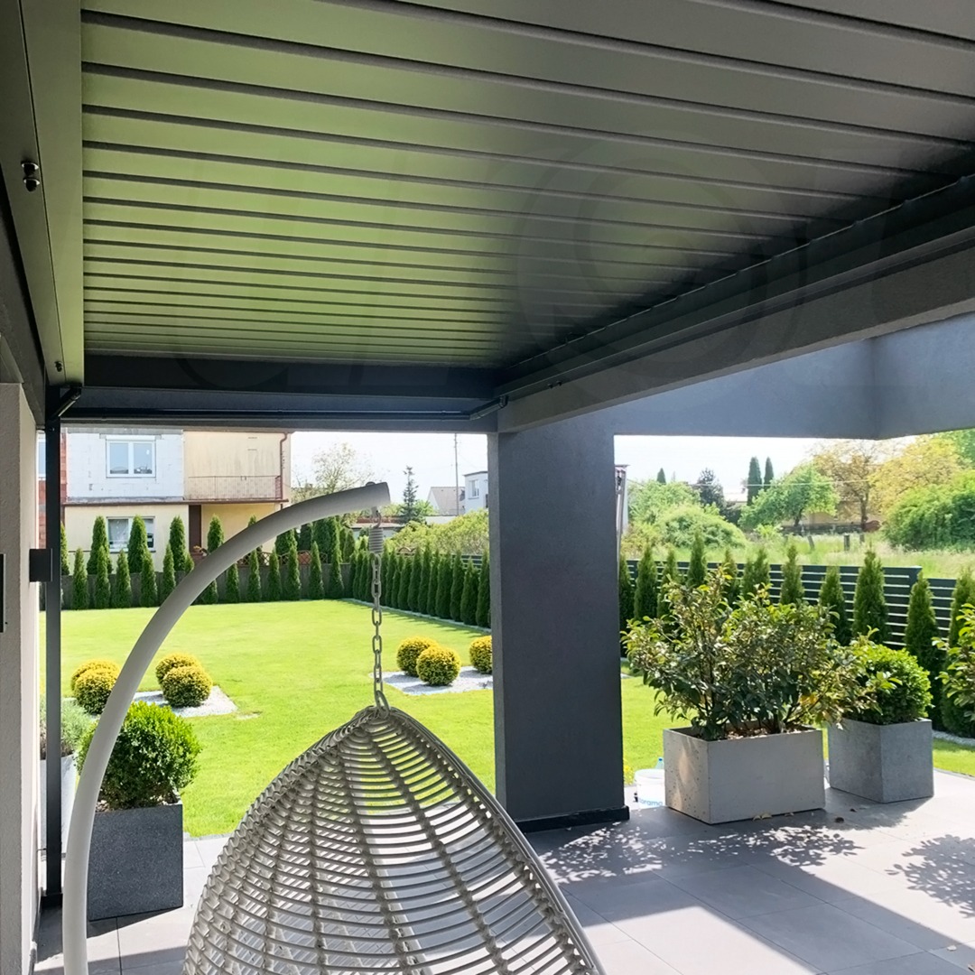 Metalowa pergola z regulowanymi lamelami zacieniająca taras z wiszącym fotelem, widok na zadbany ogród z tujami i kulistymi krzewami.