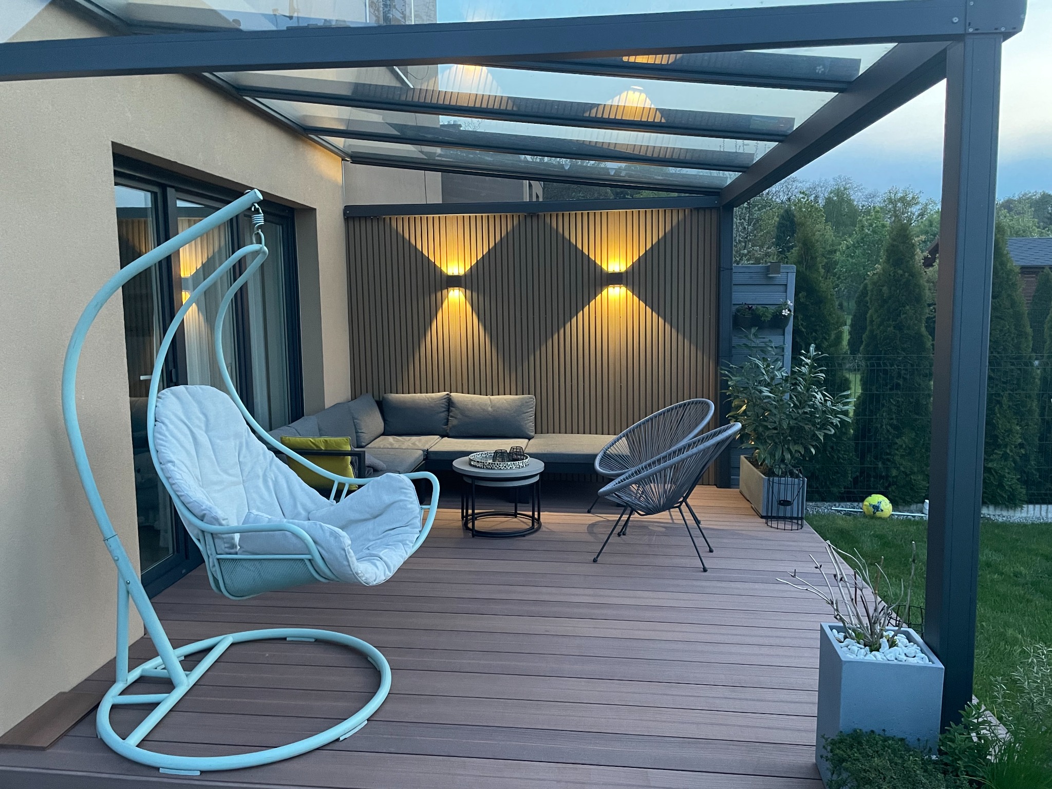 Nowoczesna przestrzeń tarasowa z zadaszeniem, oświetleniem ściennym, wiszącym fotelem i meblami ogrodowymi na drewnianej podłodze.