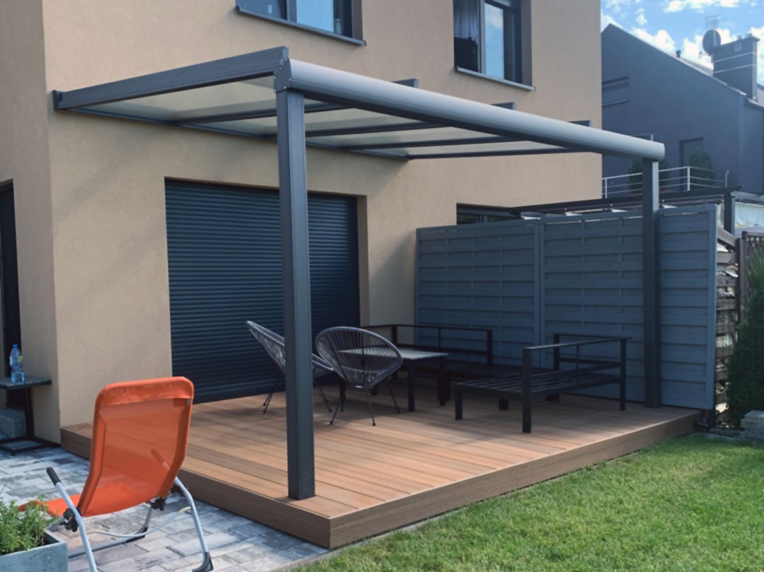Nowoczesna, metalowa konstrukcja z zadaszeniem, tworząca zacienioną przestrzeń na drewnianym tarasie przy domu. Widoczne meble ogrodowe i rolety zewnętrzne w oknie.