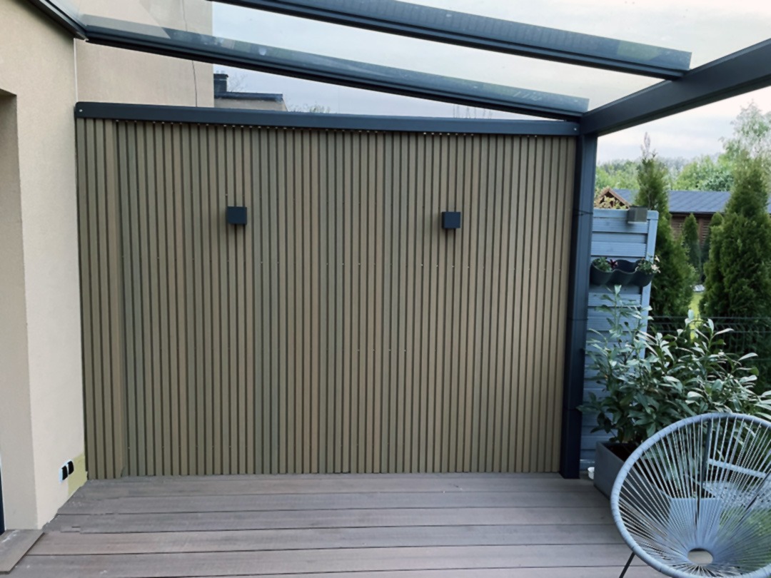 Zadaszony taras z drewnianą podłogą i pionową, drewnopodobną osłoną z dwoma lampami ściennymi, widok na ogród w tle.