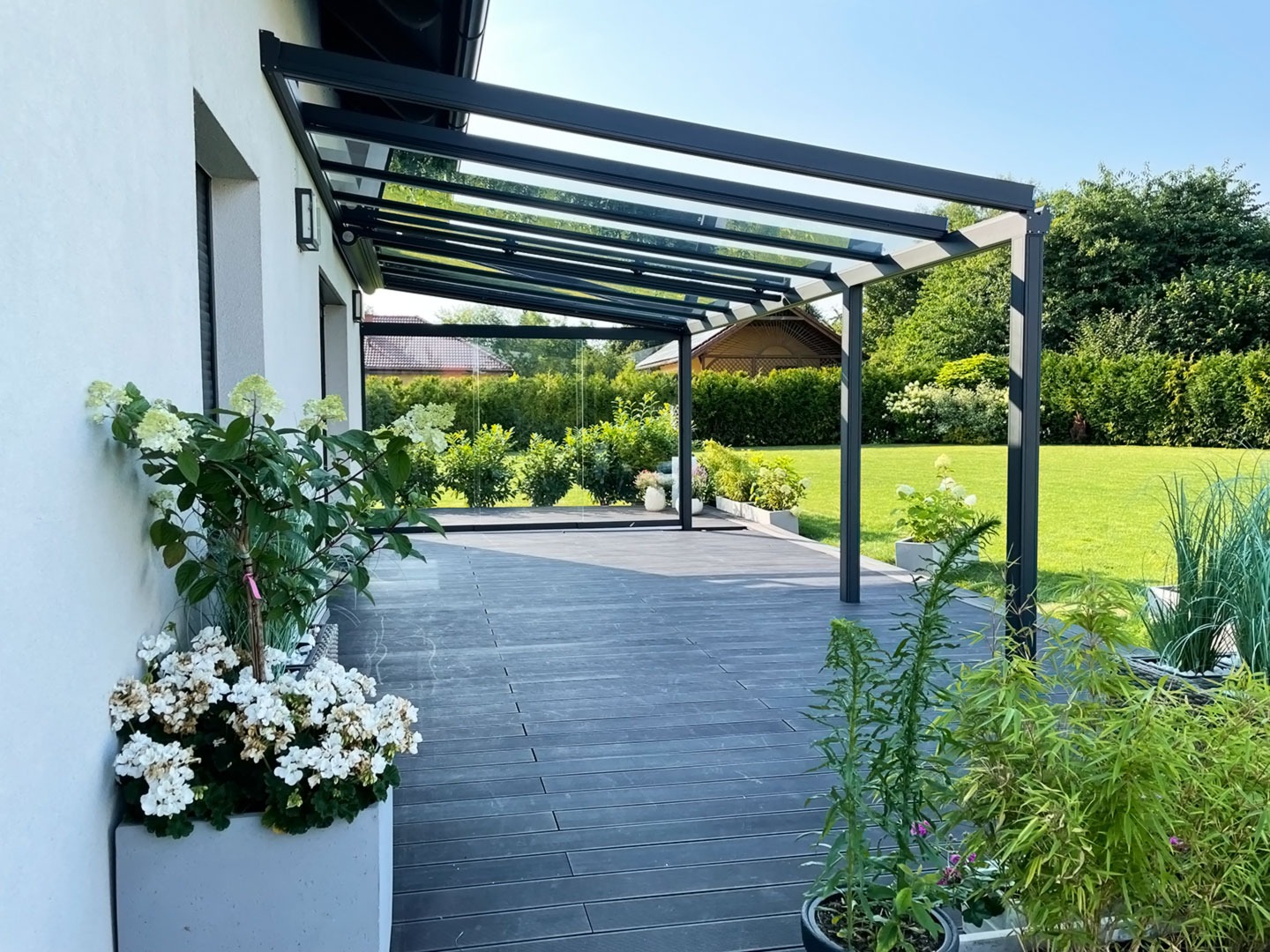 Nowoczesna, przeszklona konstrukcja z metalu, tworząca zadaszenie tarasu, otoczona zielenią i roślinami w donicach, widok na zadbany trawnik i altanę w tle.