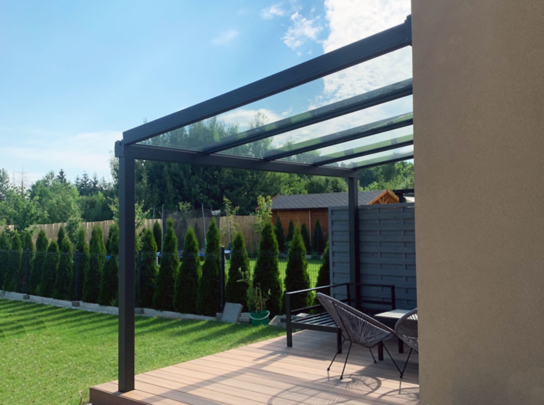 Nowoczesna pergola z metalową konstrukcją i szklanym dachem na tarasie w Opolu, widok na zadbany ogród z tujami.