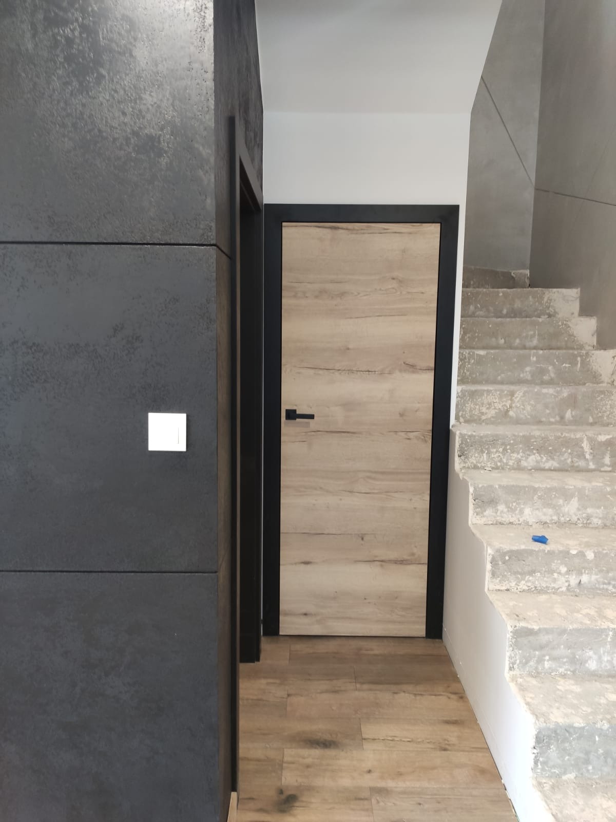 Wnętrze w trakcie wykańczania: drewnopodobne drzwi z czarną ramą i klamką, surowe betonowe schody, ciemnoszara ściana z włącznikiem, podłoga z paneli imitujących drewno.