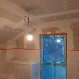 REKNES Marek Denes - Przygotowanie łazienki do prac wykończeniowych: ściany i sufit z widocznymi łatami gipsowymi, okno zabezpieczone folią, wisząca żarówka oświetla wnętrze.