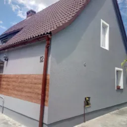 Dom jednorodzinny po remoncie elewacji, szara fasada z dekoracyjnym panelem imitującym drewno, brązowe rynny i dachówka, numer domu 12, widoczne okna i satelita.