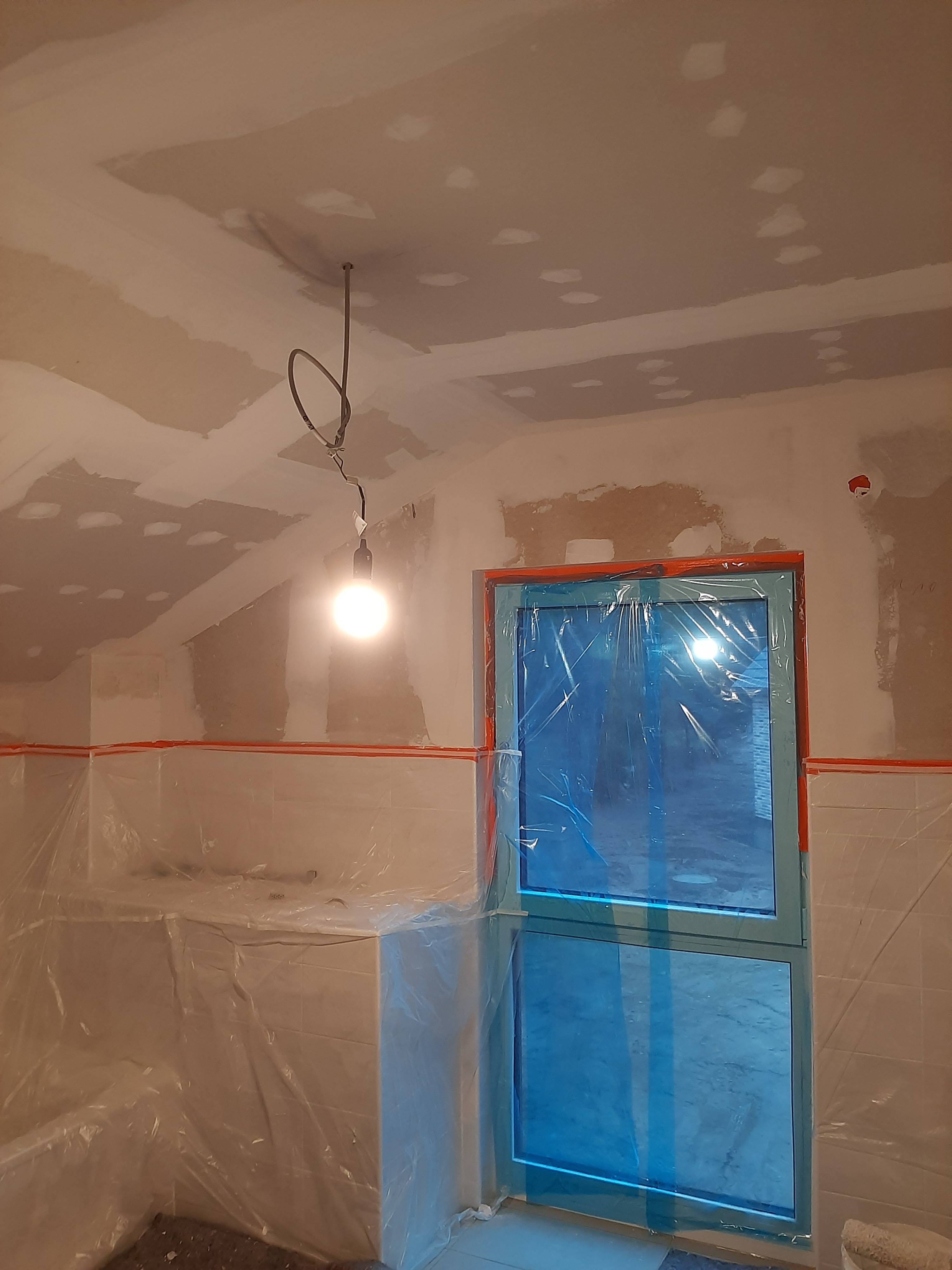 Przygotowanie łazienki do prac wykończeniowych: ściany i sufit z widocznymi łatami gipsowymi, okno zabezpieczone folią, wisząca żarówka oświetla wnętrze.