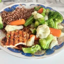 Talerz z kaszą gryczaną, grillowaną piersią kurczaka i gotowanymi warzywami: brokułami, kalafiorem i marchewką, na ozdobnym talerzu z niebieskim brzegiem.