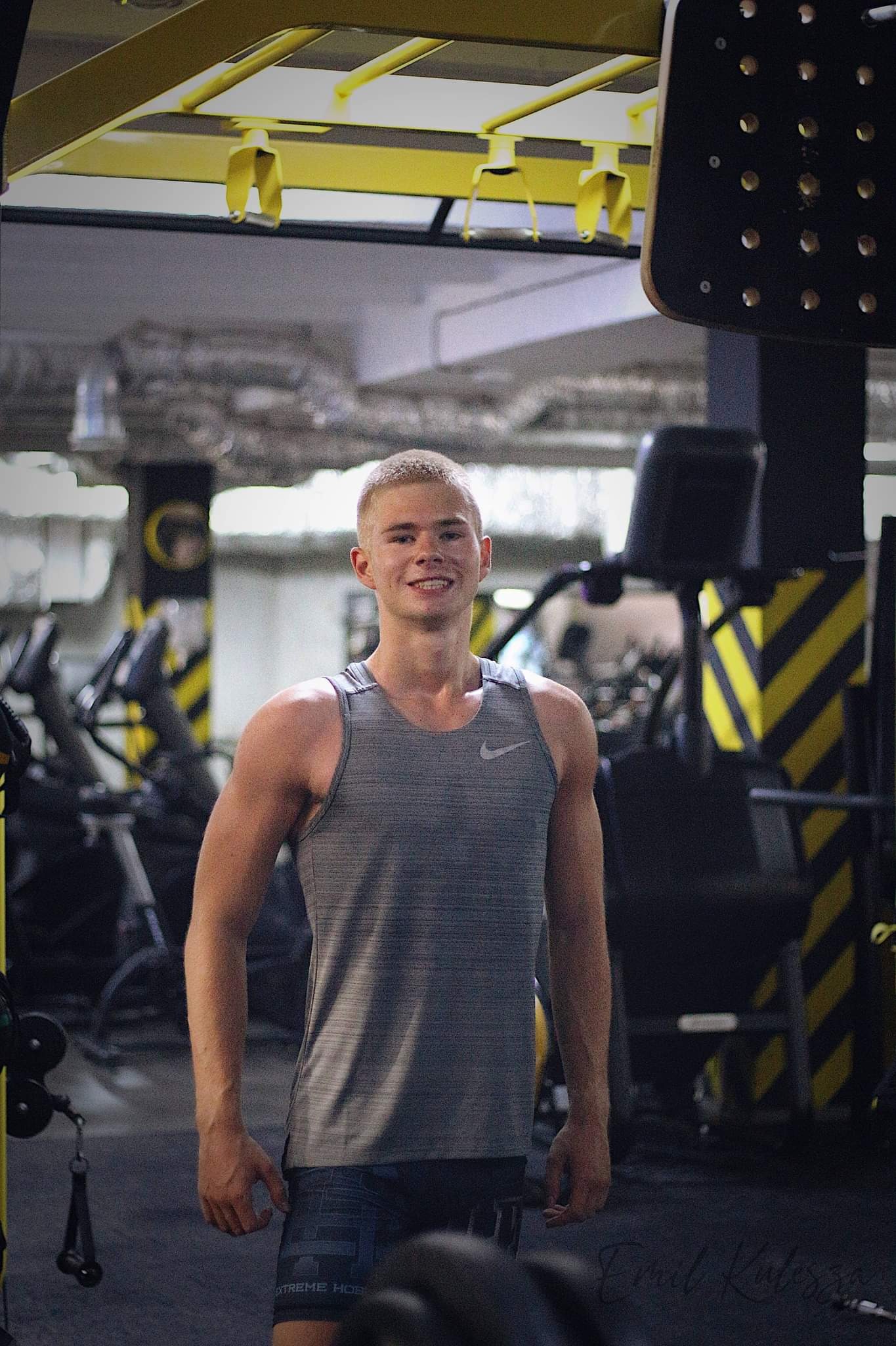 Uśmiechnięty mężczyzna w szarej koszulce bez rękawów, stojący w siłowni z żółto-czarnymi pasami ostrzegawczymi na słupie w tle, widoczne maszyny do ćwiczeń.