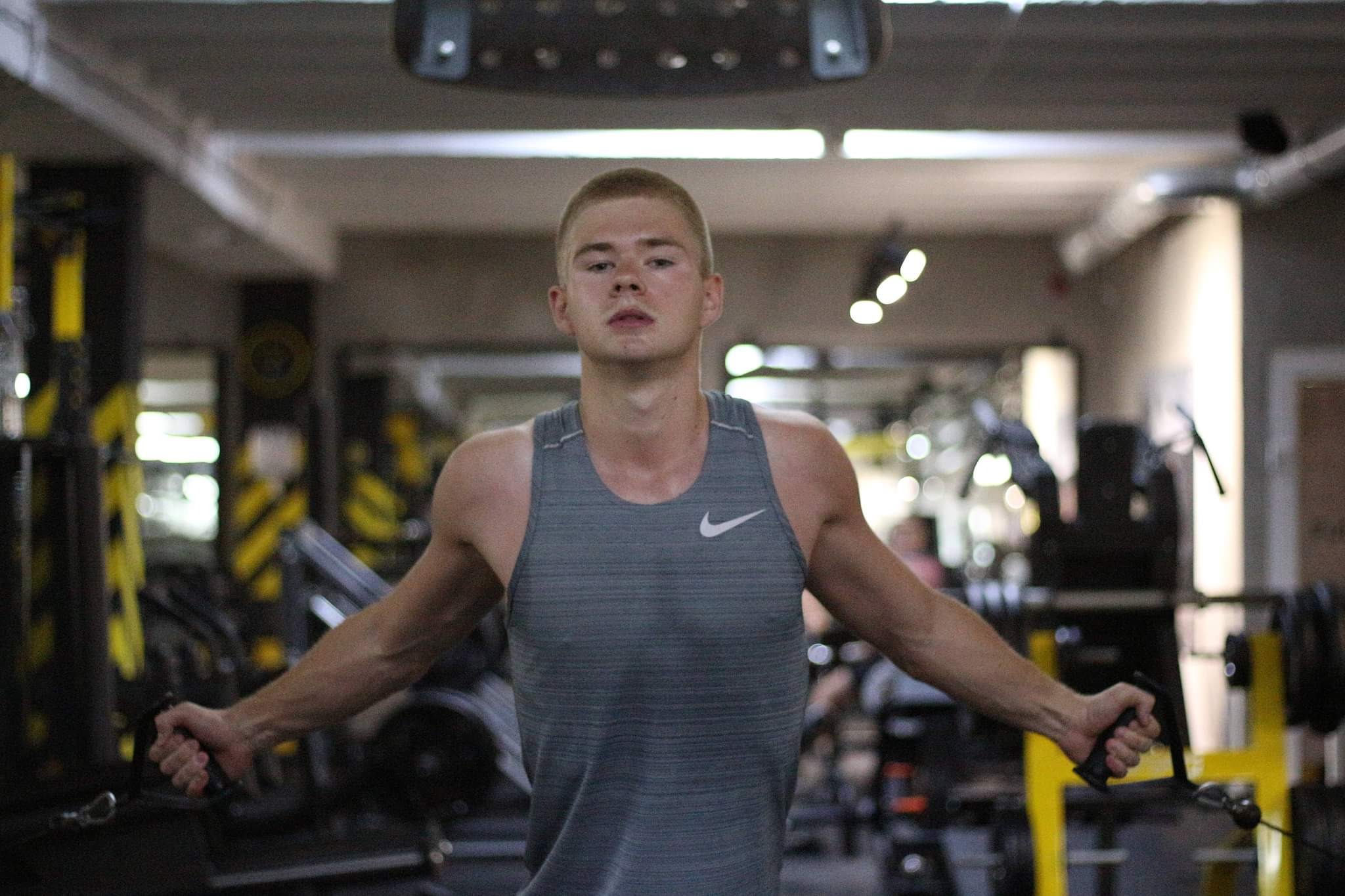 Młody mężczyzna w szarej koszulce na ramiączkach z logo Nike, ćwiczący na maszynie wyciągowej w siłowni, z widocznymi obciążeniami i żółtymi elementami konstrukcyjnymi sprzętu.
