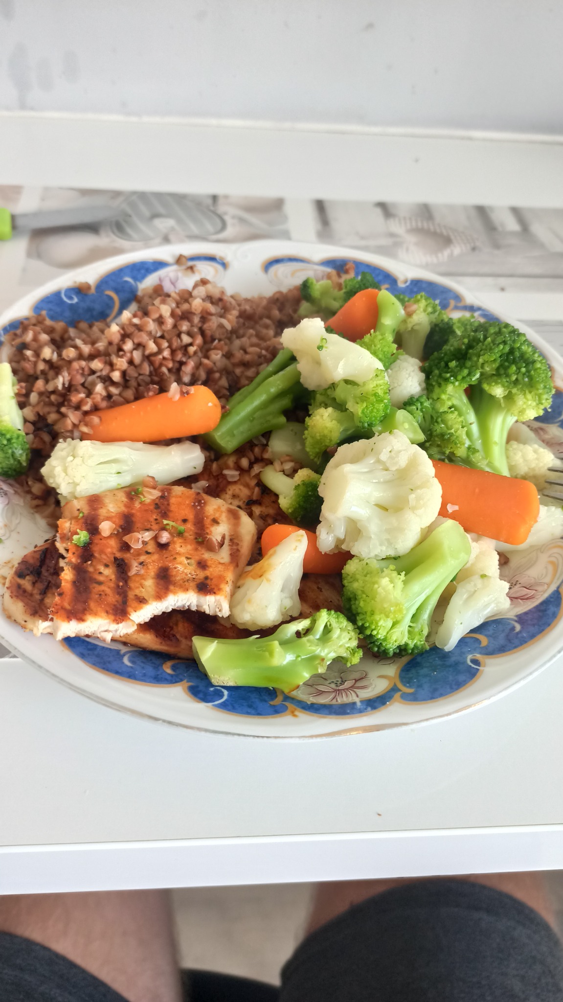 Talerz z kaszą gryczaną, grillowaną piersią kurczaka i gotowanymi warzywami: brokułami, kalafiorem i marchewką, na ozdobnym talerzu z niebieskim brzegiem.