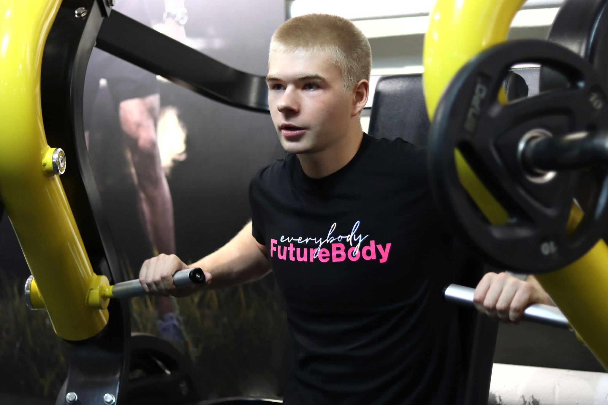 Młody mężczyzna ćwiczy na maszynie do wyciskania na klatkę piersiową w siłowni, ubrany w czarną koszulkę z napisem 'everybody FutureBody' w kolorze różowym i białym, z widocznymi obciążeniami...