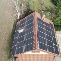 Widok z góry na dom z brązowym dachem pokrytym panelami słonecznymi; obok domu pole, drzewa i brukowany podjazd.