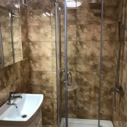Wykończona łazienka z kabiną prysznicową narożną, umywalką na szafce i brązowymi płytkami imitującymi marmur na ścianach.