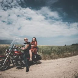 Para w średnim wieku pozuje na czerwonym motocyklu z szybą na polnej drodze pod pochmurnym niebem. Mężczyzna w czarnej koszulce z krótkim rękawem i jeansach, kobieta w kwiecistej sukience.