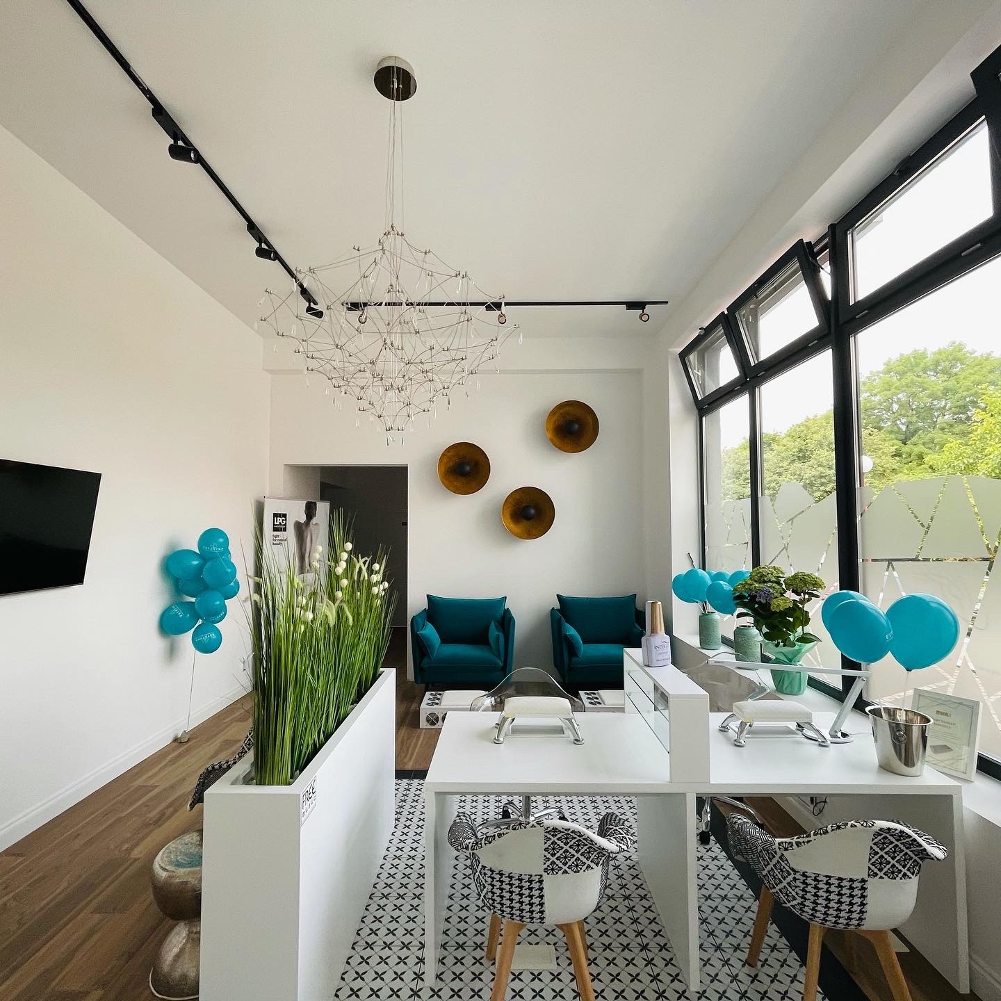 Jasne wnętrze salonu z dwoma stanowiskami do manicure, ozdobione turkusowymi balonami, nowoczesnym żyrandolem i zielonymi akcentami roślinnymi.