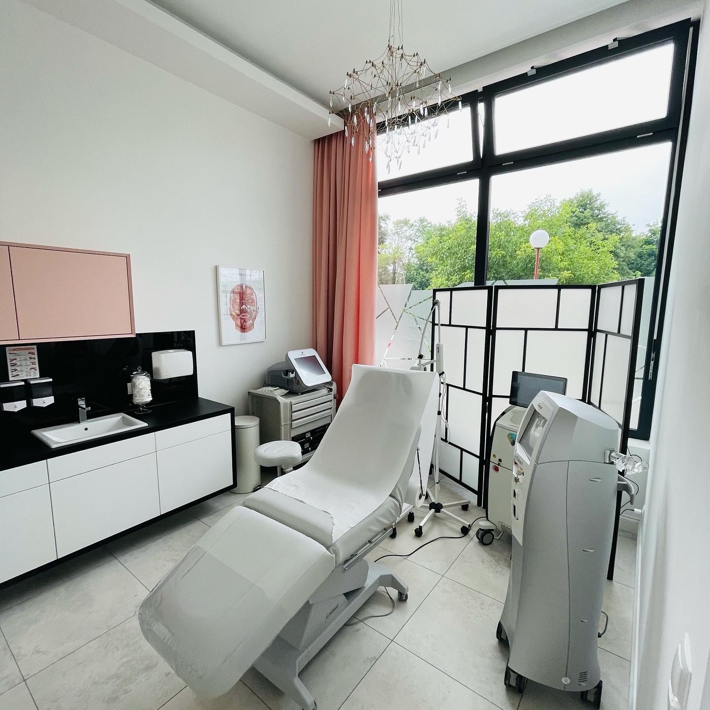 Jasne wnętrze gabinetu z białym, regulowanym fotelem zabiegowym, nowoczesnymi urządzeniami medycznymi, umywalką z czarnym blatem i dużym oknem z widokiem na zieleń, oświetlone ozdobnym żyrandolem.