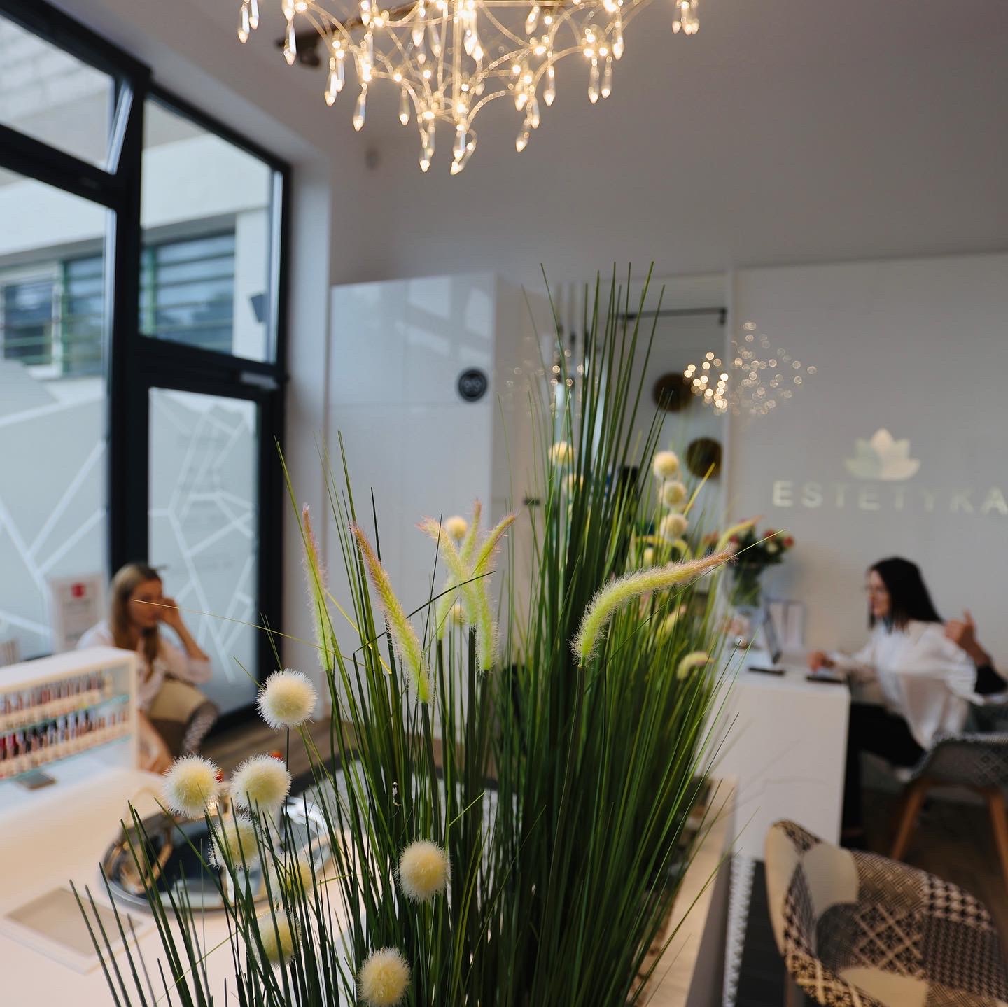 Jasne wnętrze salonu z dekoracyjną rośliną na pierwszym planie, w tle stanowiska pracy i kobiety w białych koszulach, nad głową kryształowy żyrandol, logo salonu ESTETYKA na ścianie.
