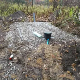 Przydomowa oczyszczalnia ścieków w trakcie budowy, widoczne rury drenażowe zasypane żwirem, czarna studzienka rewizyjna i rura wentylacyjna wystająca ponad poziom gruntu na tle drzew.