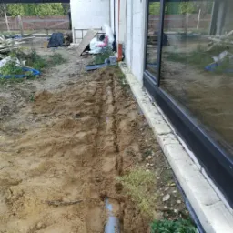 Wykop w ziemi z ułożonym szarym rurociągiem wzdłuż ściany budynku z dużym oknem; widoczne ślady stóp w wykopie i elementy instalacji na zewnątrz.
