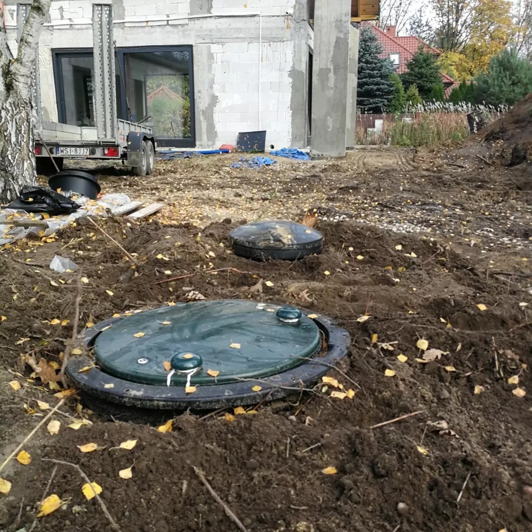 Dwa zielone włazy do podziemnych zbiorników w wykopie ziemnym na tle budowy domu w stanie surowym otwartym i przyczepy.