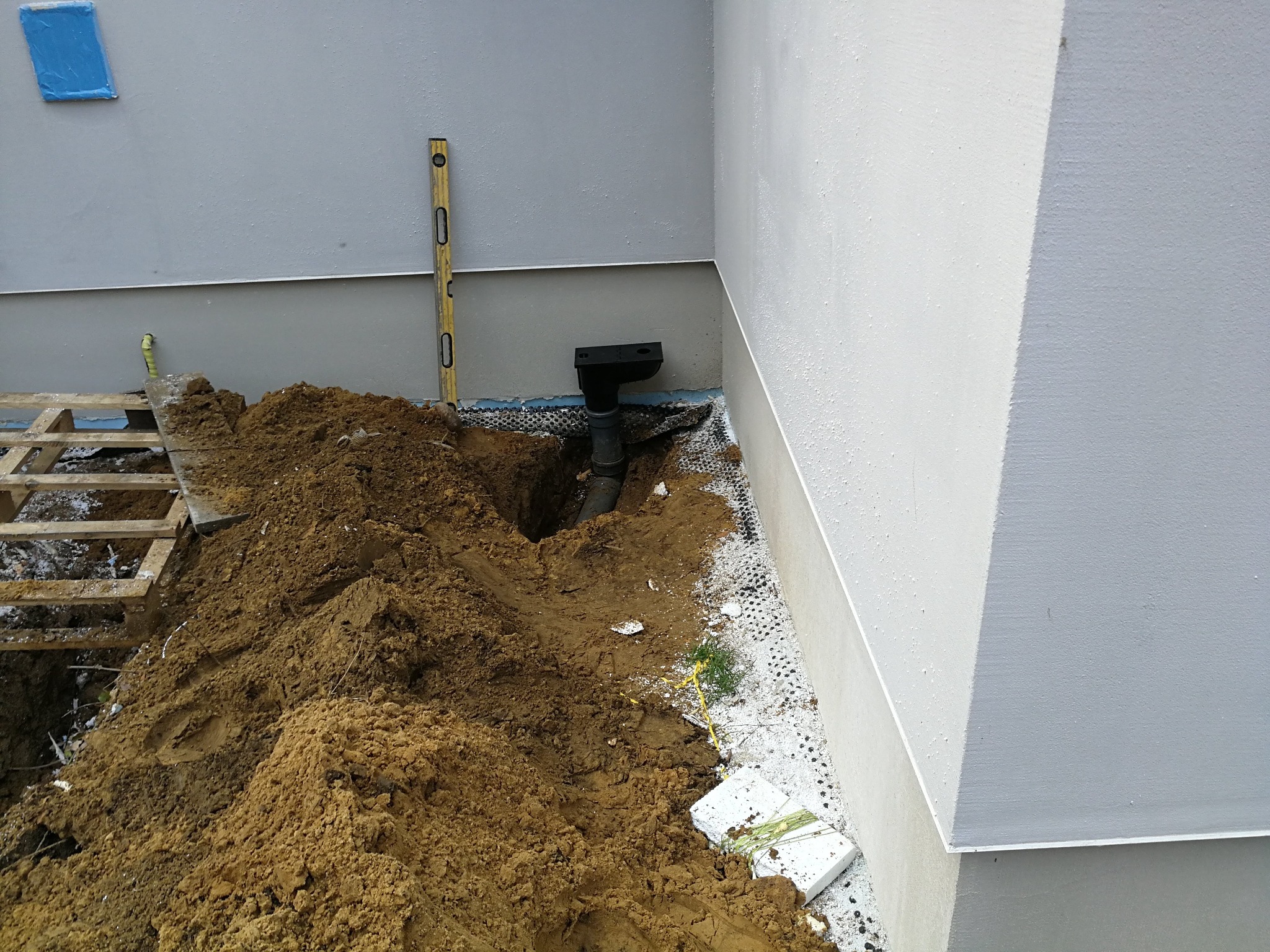 Wykopany rów przy fundamencie budynku z widoczną rurą kanalizacyjną i poziomicą opartą o ścianę.