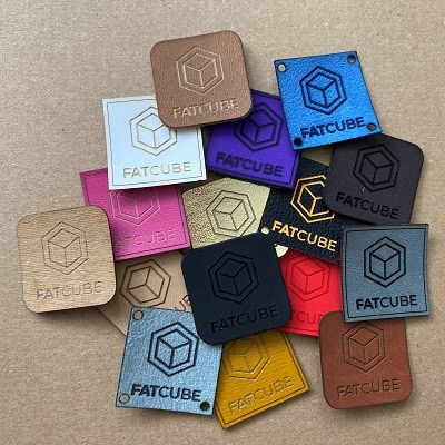 Kolekcja skórzanych i materiałowych etykiet z zaokrąglonymi rogami i wygrawerowanym logo 'FATCUBE' w różnych kolorach i fakturach ułożonych na beżowym tle.