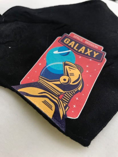 Czarna tkanina z naklejoną kolorową grafiką przedstawiającą astronautę i napis 'SPACE MISSION GALAXY' na czerwonym tle.