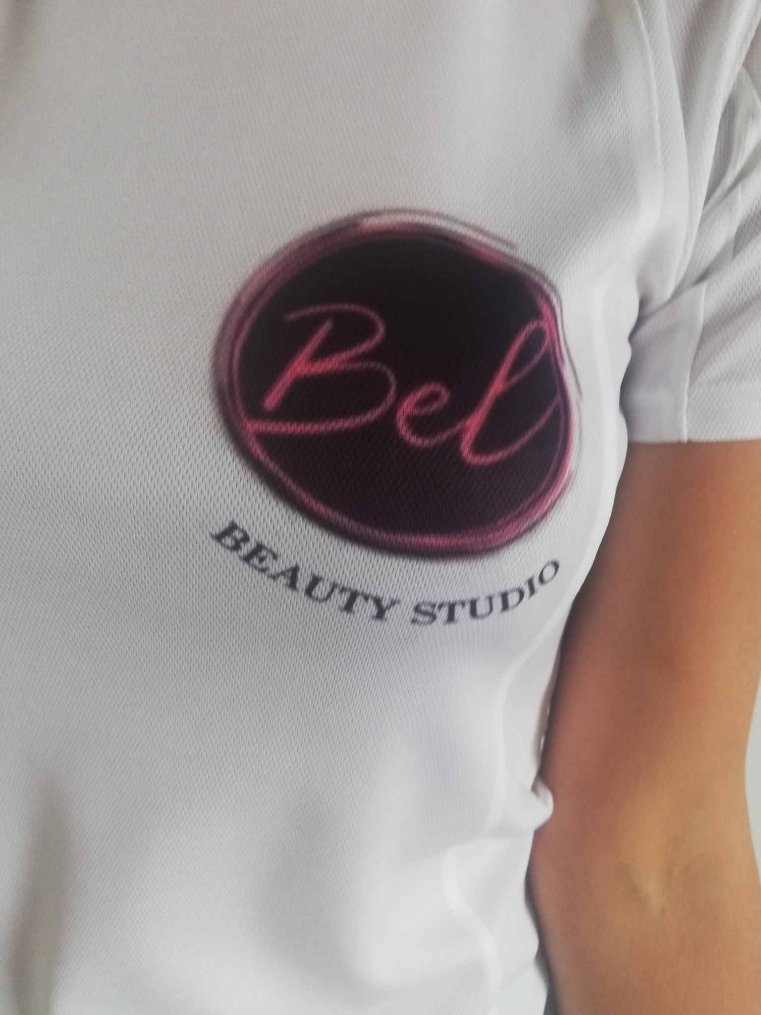Biała koszulka z siateczki z nadrukowanym logo 'Bel Beauty Studio' w okrągłej ramce z różowym napisem i obwódką.