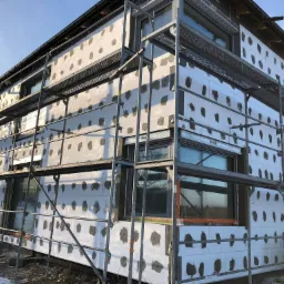 Dwukondygnacyjny dom w trakcie montażu białych płyt izolacyjnych na elewacji, widoczne szare plamy kleju i metalowe rusztowanie na tle błękitnego nieba.