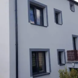 Świeżo pomalowana elewacja budynku z szarymi obramowaniami okien i ciemną rynną, widok z dołu na fragment ściany i balkon.