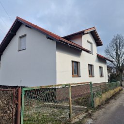 Paweł Porębski Natalia Kalczuk - Świeżo otynkowany, biały dom jednorodzinny z brązową dachówką, widziany zza zielonego, metalowego ogrodzenia w pochmurny dzień.