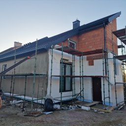 Paweł Porębski Natalia Kalczuk - Dom w trakcie prac budowlanych, widoczne rusztowania, ocieplenie styropianem na elewacji oraz nowy, czarny dach z kominem. Część elewacji w stanie surowym z czerwonej cegły. Na pierwszym planie...