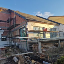 Paweł Porębski Natalia Kalczuk - Dom w trakcie prac budowlanych: część elewacji ocieplona styropianem, część w surowej cegle, rusztowania wokół budynku, przygotowane płyty izolacyjne, niezagospodarowany teren wokół.