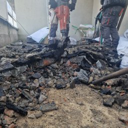 Paweł Porębski Natalia Kalczuk - Demontaż starego pokrycia dachowego za pomocą młota pneumatycznego, widoczne gruz, resztki papy i fragment drabiny oparta o ścianę budynku.