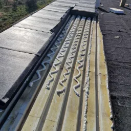 Widok z góry na fragment dachu w trakcie naprawy, z widocznymi warstwami izolacji i świeżo nałożoną pianką uszczelniającą w zygzakowatych liniach między panelami, z fragmentem starego pokrycia po...