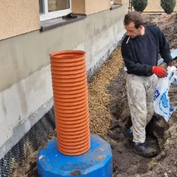 Prace ziemne przy izolacji fundamentów: robotnik z papierosem w ustach, w ubrudzonych farbą spodniach i czerwonych rękawicach, trzyma worek z gruzem obok pomarańczowej rury kanalizacyjnej...