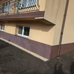 Fragment budynku z otynkowaną elewacją w odcieniach beżu i brązu, z widocznym balkonem z ozdobną, metalową balustradą i świeżo obsypanym kamieniami fundamentem.