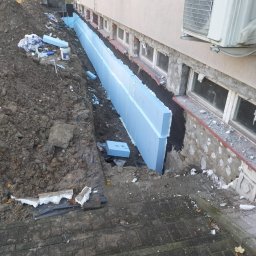 Paweł Porębski Natalia Kalczuk - Izolacja fundamentów budynku mieszkalnego z użyciem niebieskiego styropianu. Wykop wzdłuż ściany, widoczne okna piwniczne i klimatyzator na elewacji.