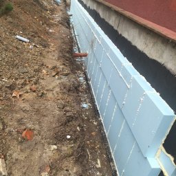 Paweł Porębski Natalia Kalczuk - Izolacja fundamentów budynku z użyciem niebieskiego styropianu, zabezpieczonego czarną folią hydroizolacyjną. Widoczny wykop i rura drenażowa.