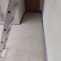 Paweł Porębski Natalia Kalczuk - Przygotowanie do izolacji fundamentów: widoczna ściana, podłoga z płytkami i drabina oparta o ścianę. Czarny pas izolacyjny na styku ściany i podłogi.