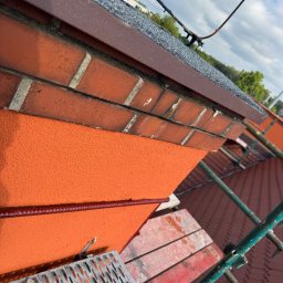 Paweł Porębski Natalia Kalczuk - Fragment elewacji z ceglanym wykończeniem i pomarańczowym tynkiem strukturalnym, widok z rusztowania na tle dachu pokrytego dachówką. Detal wykończenia.