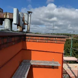 Pomarańczowa elewacja budynku z ceglanym wykończeniem dachu, widoczne elementy instalacji i rusztowania na tle błękitnego nieba z chmurami.