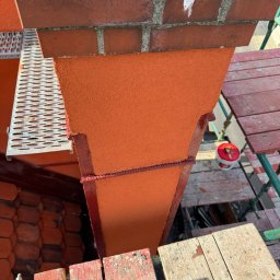 Paweł Porębski Natalia Kalczuk - Ocieplony komin z rusztowania, wykończony pomarańczowym tynkiem strukturalnym, z widocznymi elementami ceglanymi i narzędziami malarskimi.