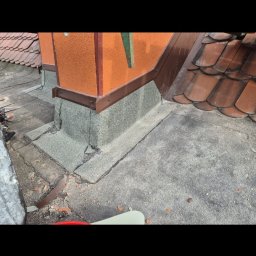 Paweł Porębski Natalia Kalczuk - Szczegółowe ujęcie komina na dachu z widocznymi elementami obróbki blacharskiej i uszczelnienia. Widoczne płytki dachowe i fragment dachu.