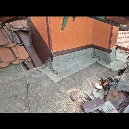 Paweł Porębski Natalia Kalczuk - Fragment dachu z kominem po naprawie, widoczne nowe obróbki blacharskie i uszczelnienia. Część starego pokrycia leży obok komina. Pomarańczowy komin z brązowymi detalami.